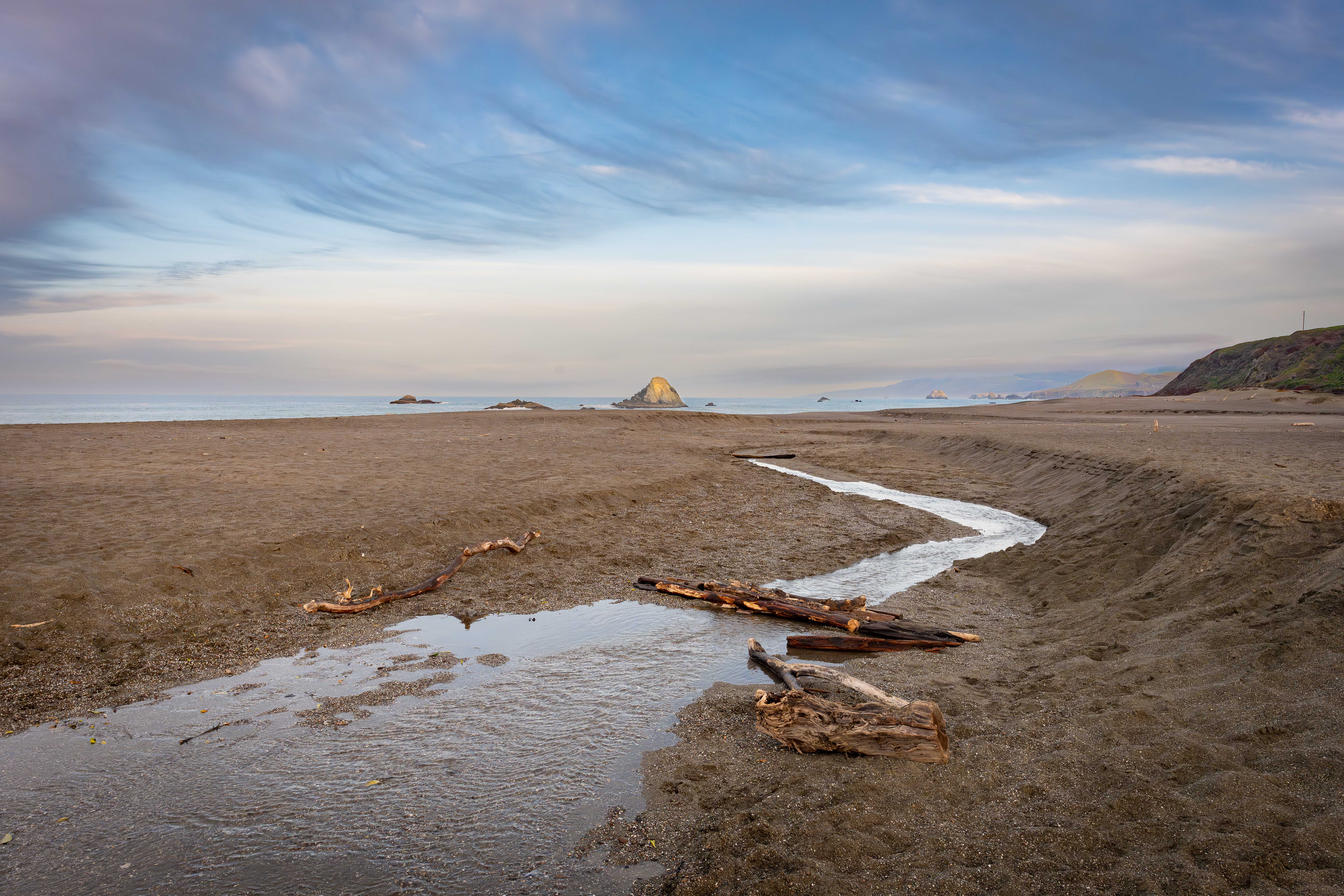 Wright's Beach on the Sonoma coast at dusk, a creek winding across the wide sand to the sea with driftwood and sea stacks